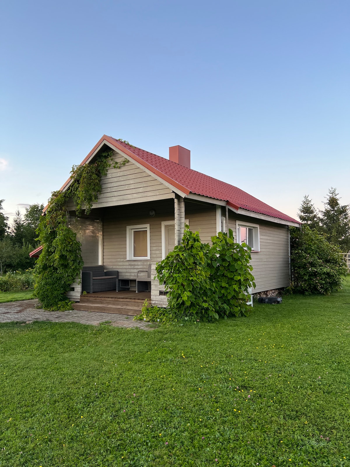 A charming two-storey country house is surrounded by lush greenery, featuring ivy-covered walls and a sloped red roof. A patio area is visible with outdoor seating, providing a welcoming space for relaxation in the natural setting.