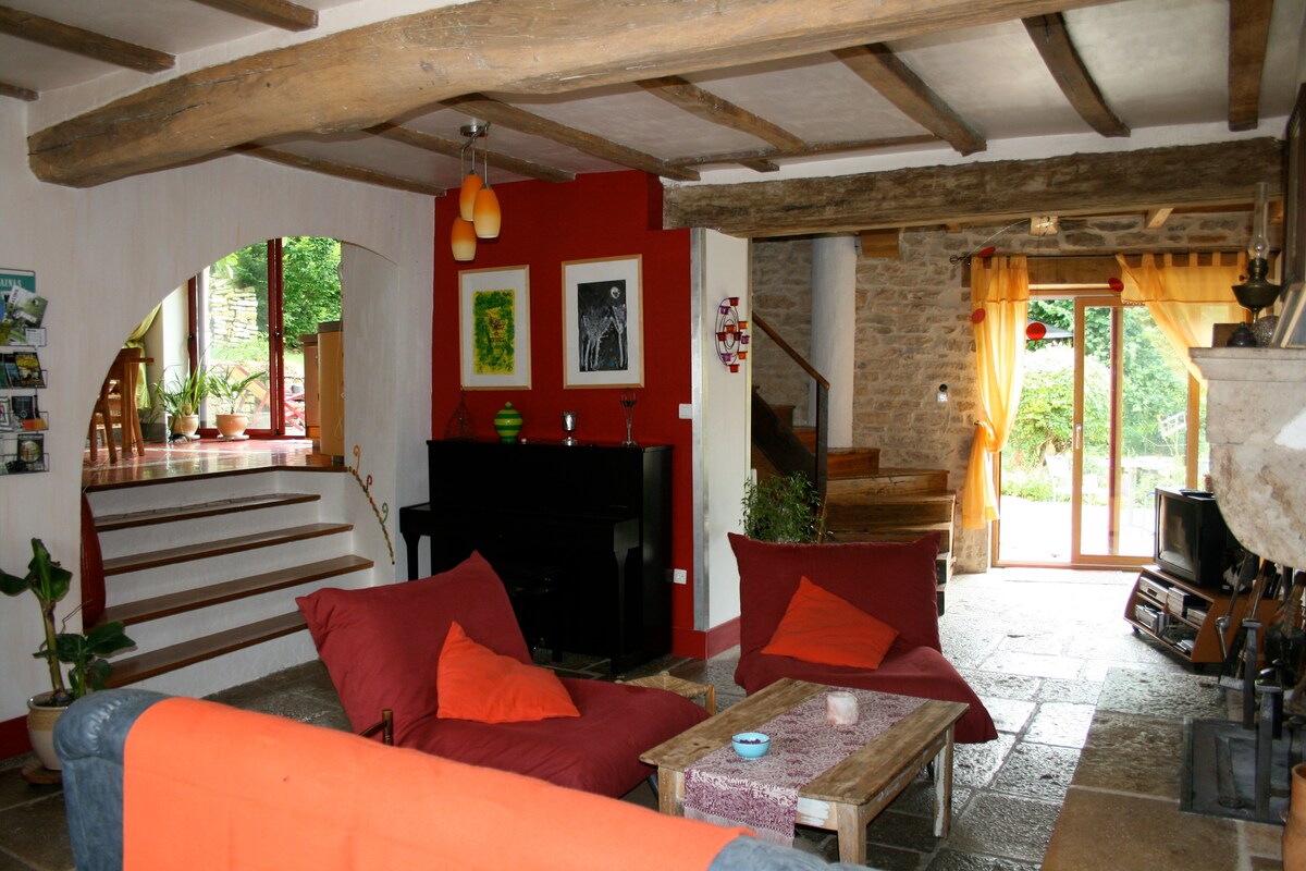 The living area features a mix of natural materials with wooden beams and stone walls. A black piano sits next to a warm red accent wall, while bold cushions are arranged on a casual seating area. Large doors lead to a lush garden outside.