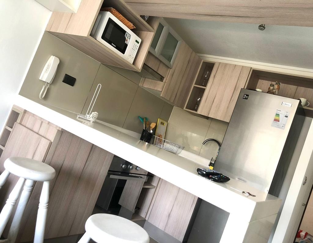 A modern kitchen features light wood cabinetry and a white countertop. Essential appliances, including a microwave and refrigerator, are visible. The design includes a sleek stovetop and open shelving for utensils. Two white bar stools are positioned at the kitchen island.