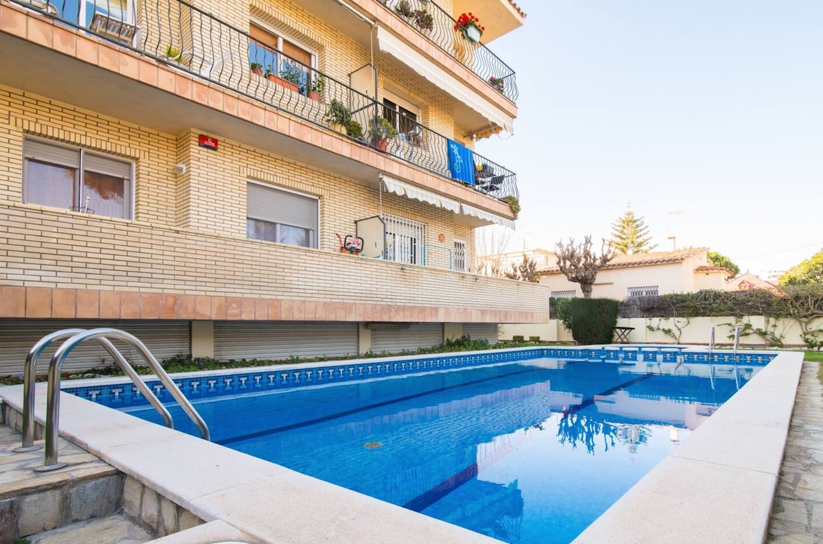 A large outdoor swimming pool is situated in front of a multi-story building, reflecting the clear blue sky. Surrounding gardens and patio areas add greenery, with residential balconies above adorned with potted plants.