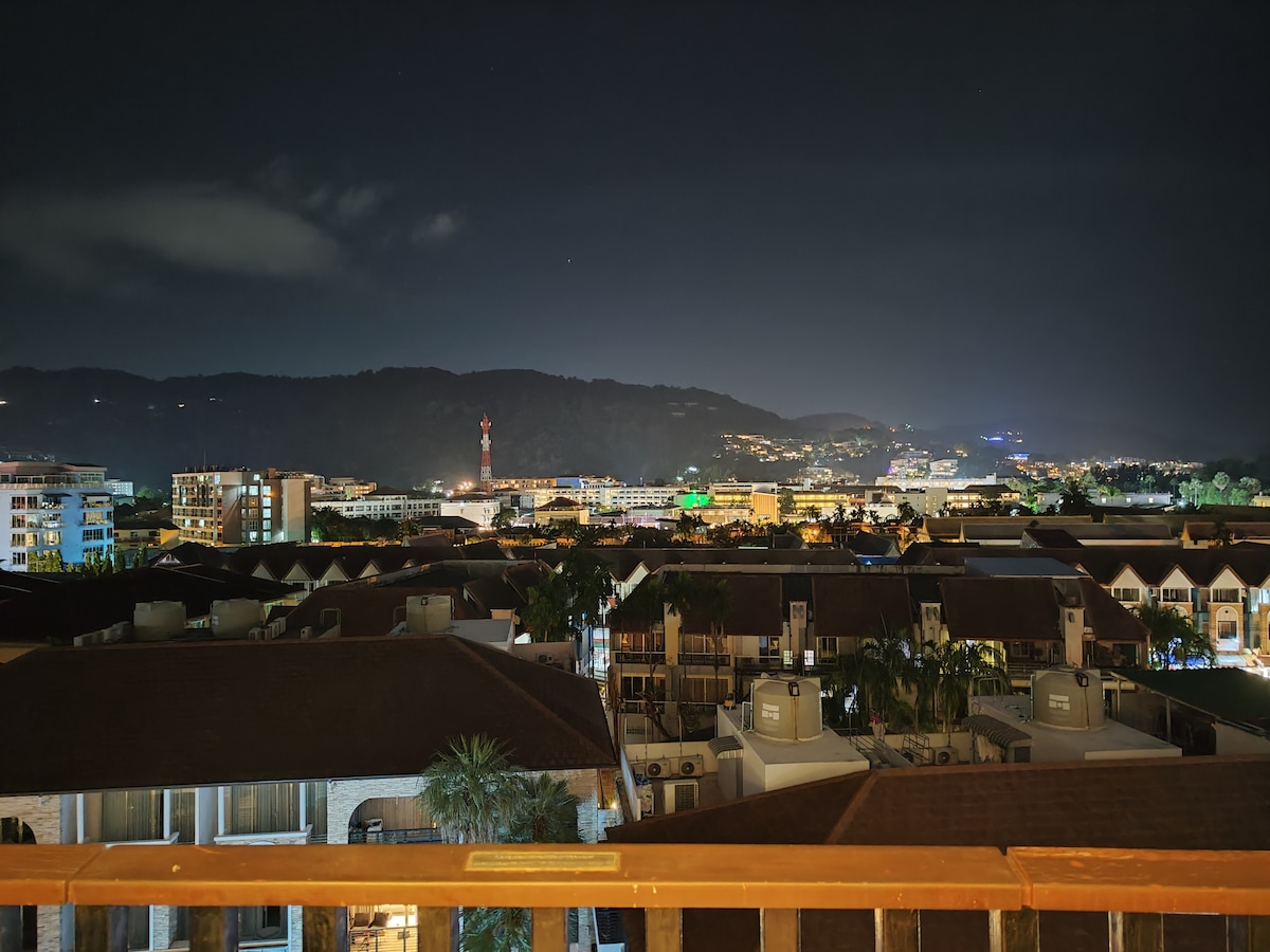 A panoramic night view displays a vibrant skyline with illuminated buildings and distant hills under a dark sky. The foreground reveals rooftops and palm trees, while various city lights create a twinkling effect across the landscape.