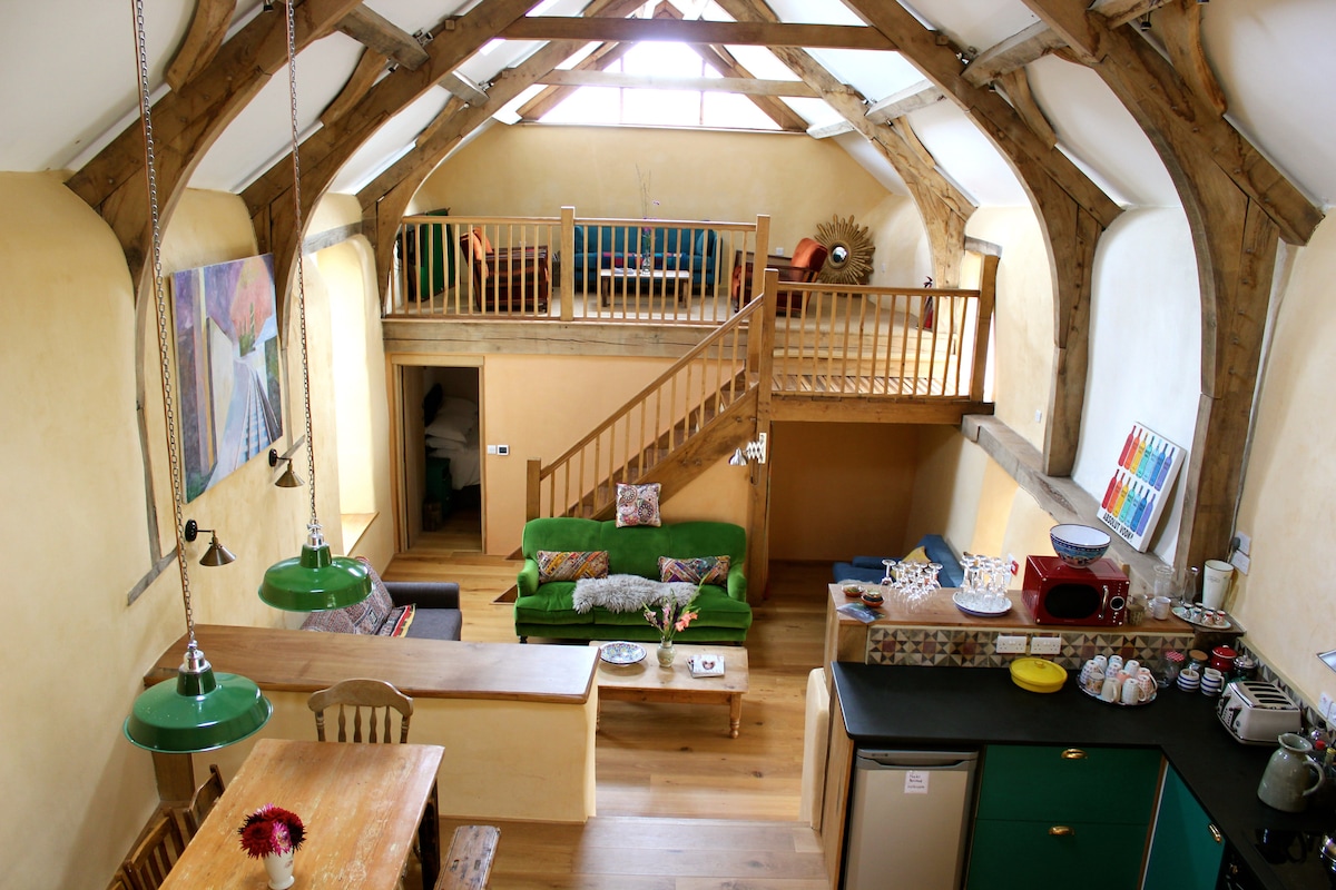 An open-plan living space showcases natural wood beams and warm, earthy walls. A spacious green sofa is complemented by decorative cushions, while a wooden dining table is set for shared meals. A loft area with seating is visible above, offering additional relaxation space.