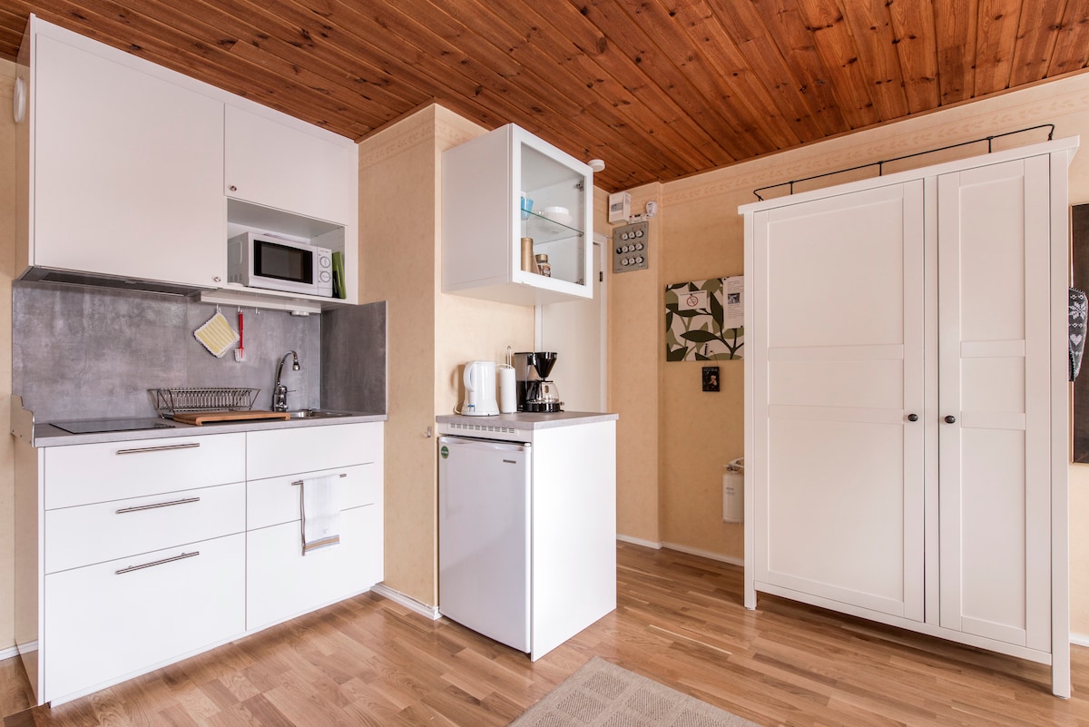 A compact kitchen area features modern white cabinets and a countertop. Included are a sink, microwave, coffee maker, and a small refrigerator. A wooden ceiling overhead provides a warm contrast to the light-colored walls and flooring, enhancing the room's simplicity and functionality.