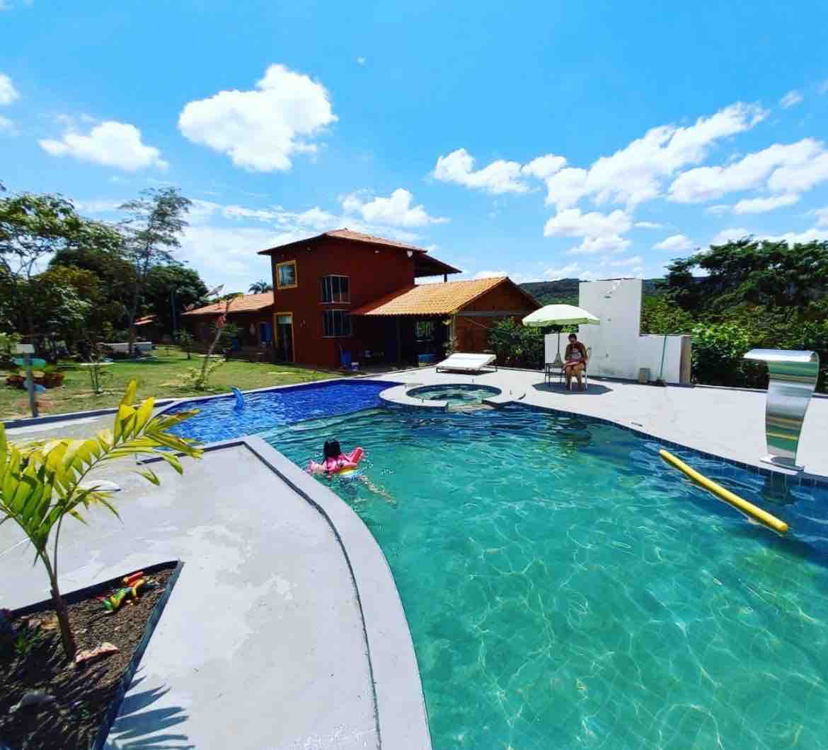 A sunny outdoor space features a refreshing swimming pool with a shallow area for relaxation. Surrounding greenery is complemented by a spacious patio area equipped with lounge chairs and umbrellas. A colorful two-story building is visible in the background, providing a welcoming atmosphere.