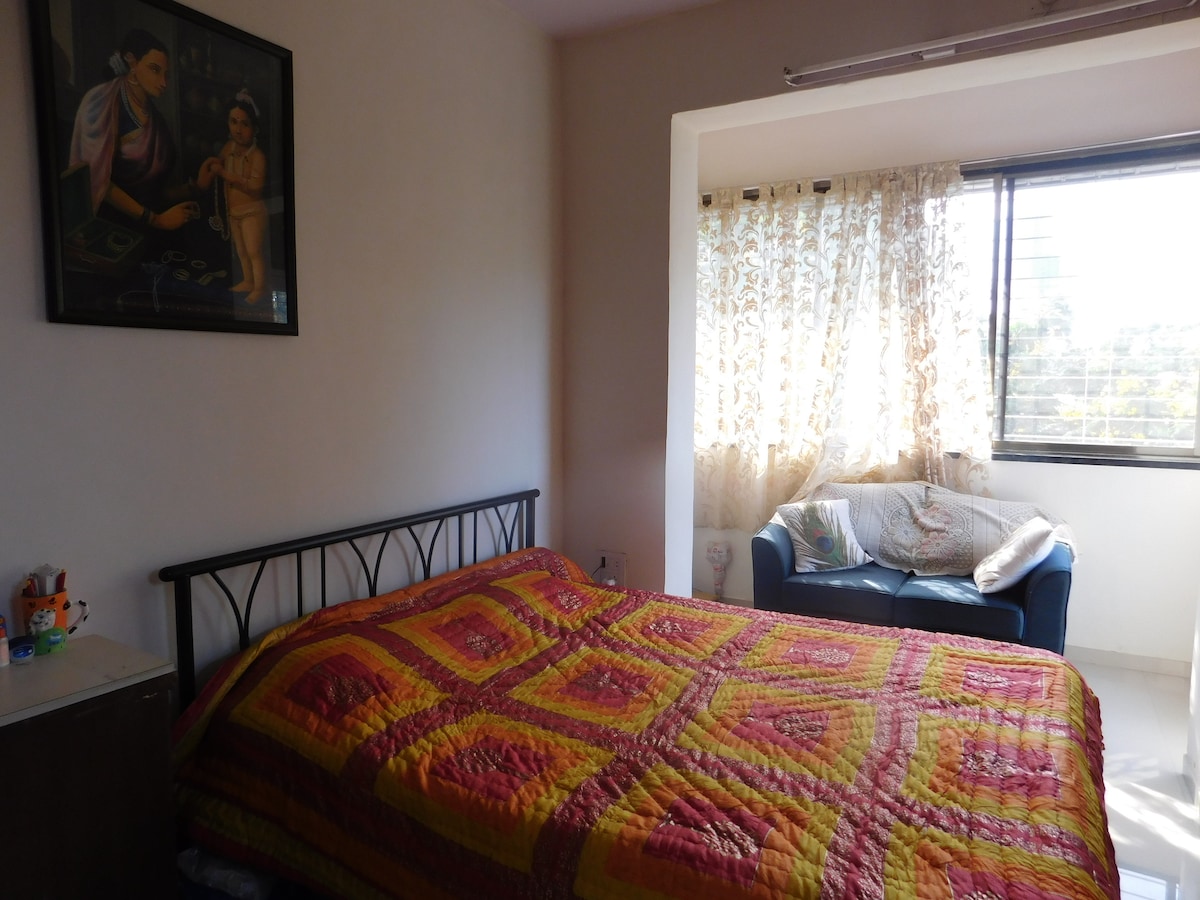 A cozy bedroom is presented with a spacious bed covered in a vibrant patterned quilt. A comfortable seating area is visible beside a large window, with sheer curtains allowing natural light to fill the room. A wall painting adds a decorative touch.