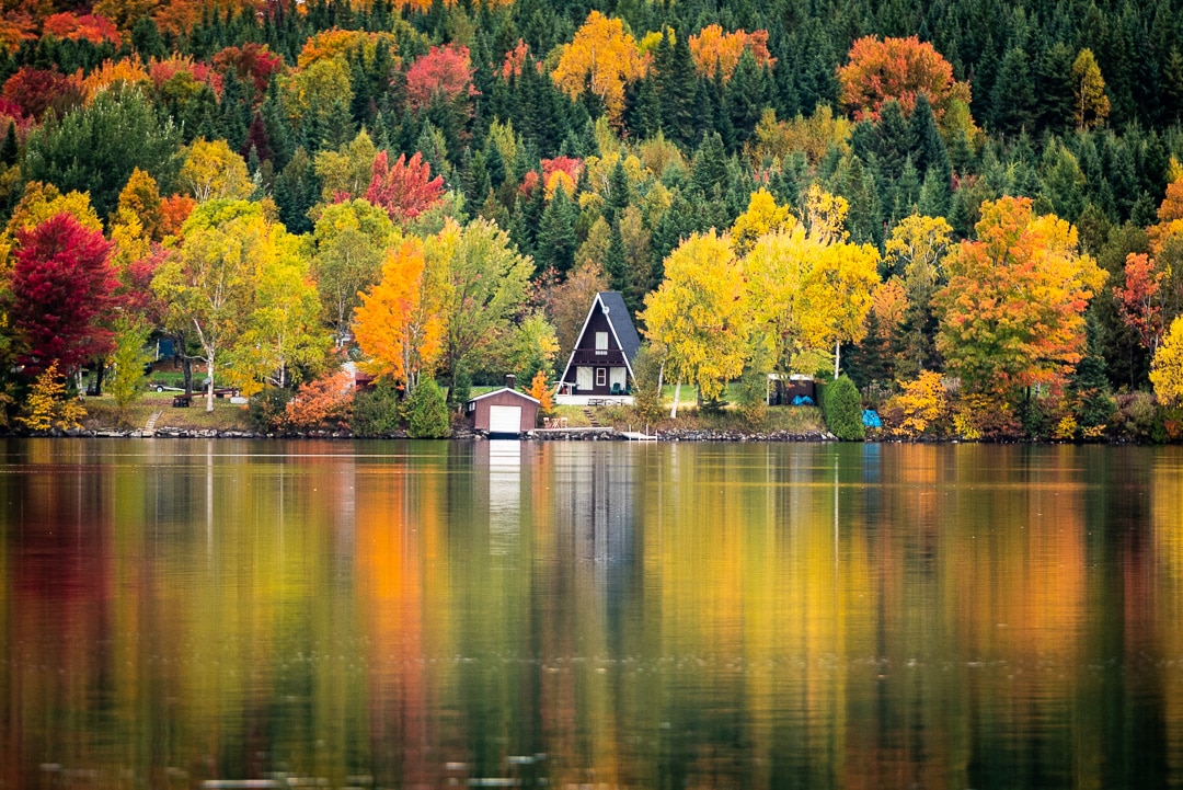 An A-frame chalet is situated by the lake, surrounded by vibrant autumn foliage. The water reflects the colorful trees, creating a serene and harmonious view. A boathouse can be seen at the water's edge, enhancing the peaceful lakeside atmosphere.