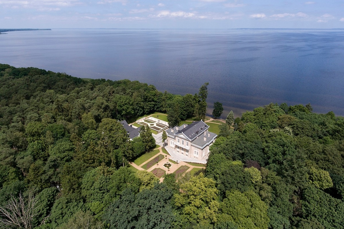 Aerial view of the historic property surrounded by lush greenery and located near the bay. The building's architecture complements the landscape, with expansive grounds leading to the water's edge. The calm water reflects the sky, creating a serene atmosphere.