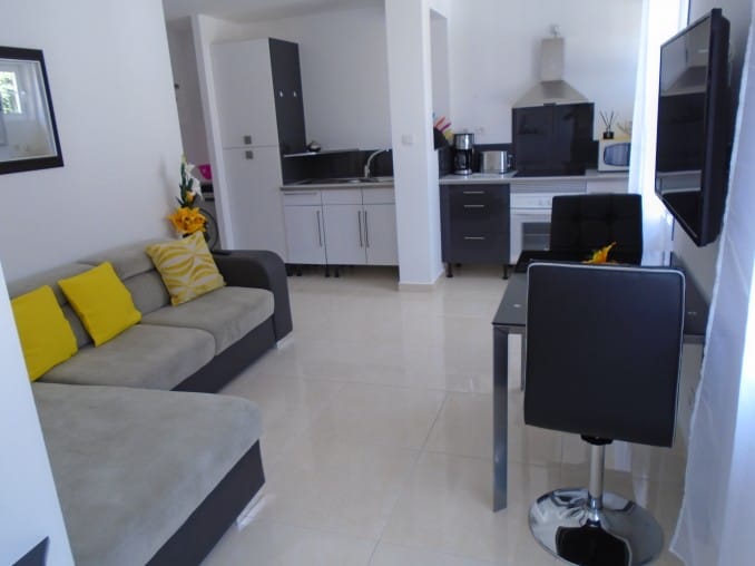 A modern living area features a gray sofa adorned with bright yellow cushions. A sleek dining table with black chairs complements the space. The kitchen area is equipped with contemporary appliances, including a microwave and stovetop, all set against clean, white walls and shiny tiled floors.