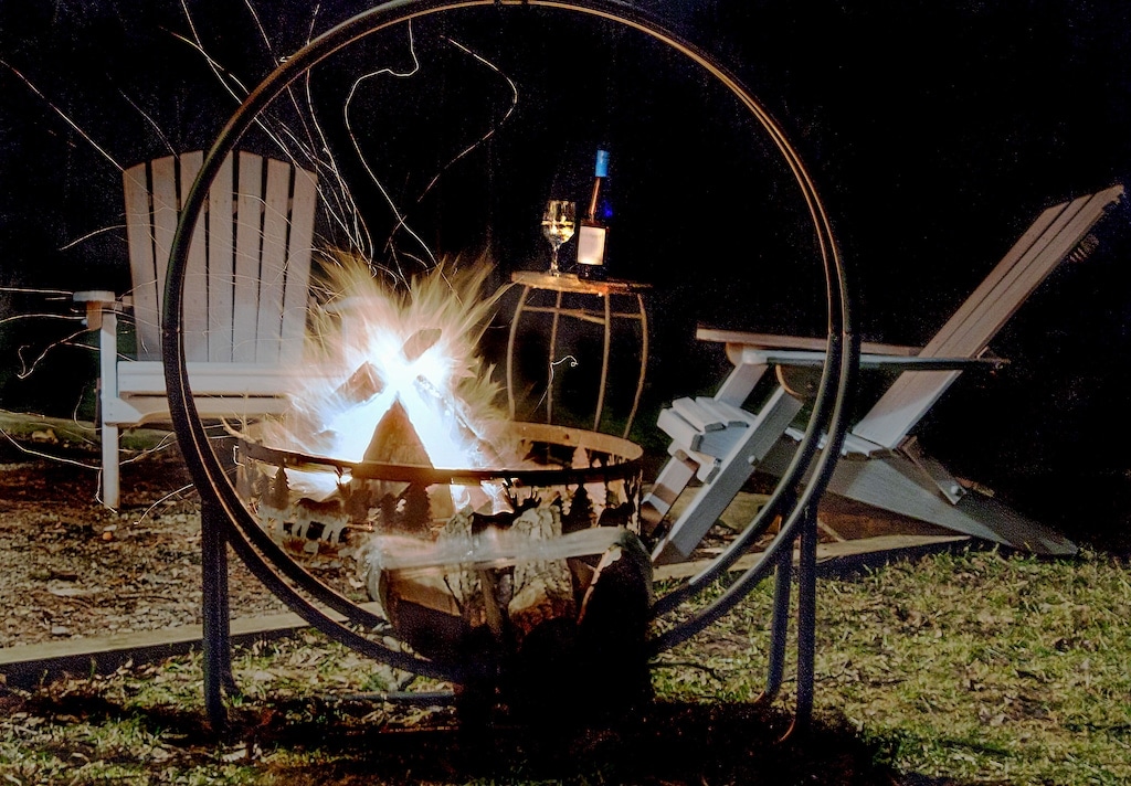 A circular fire pit is positioned prominently, with flames brightly erupting from the center. Two outdoor chairs are arranged nearby, and a small table holds a bottle of wine and a glass, creating a relaxed outdoor setting for gatherings.