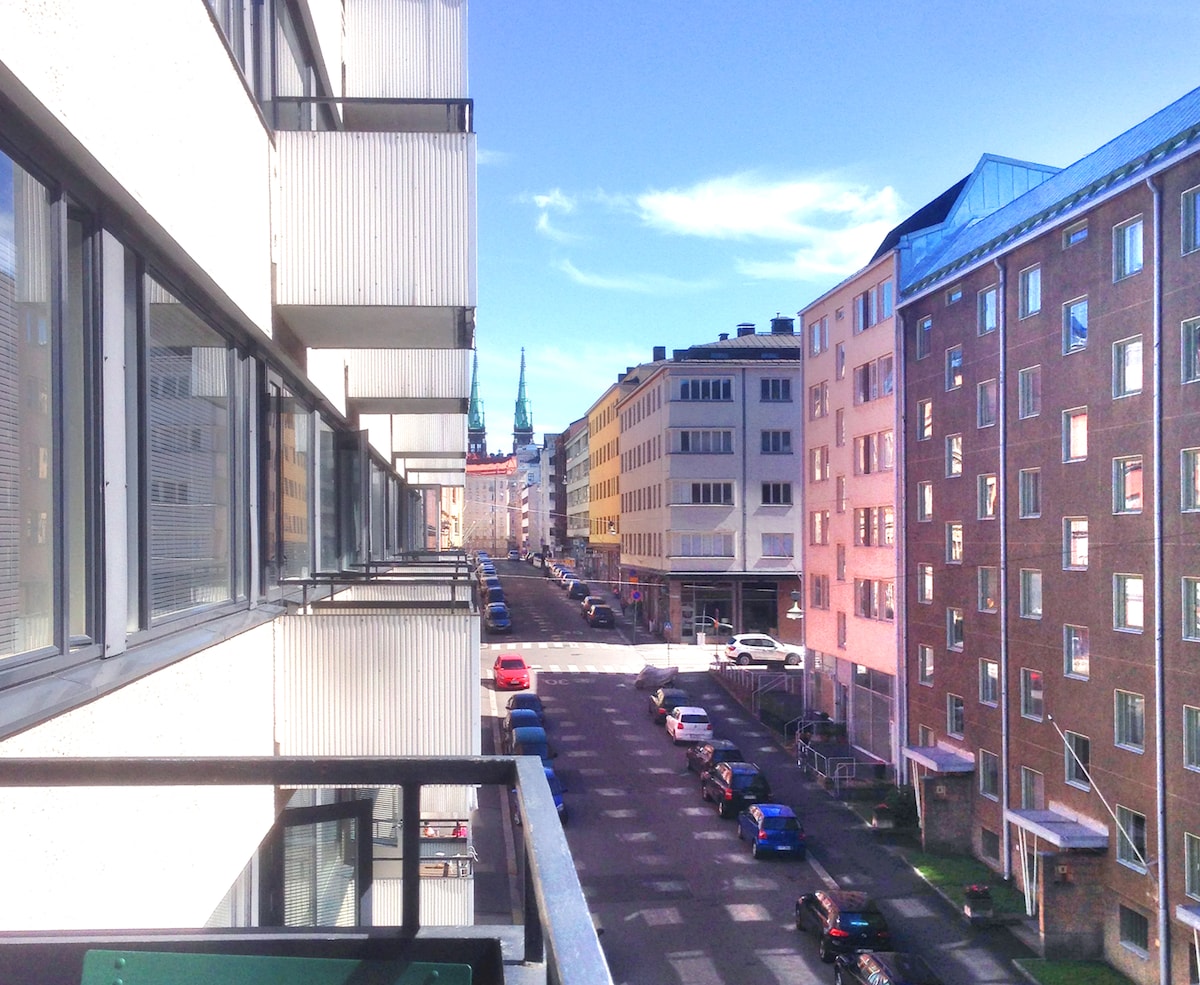 A view from the balcony captures a sunny street scene with a mix of modern and classic architectural styles. Cars are parked along the road, and the distant skyline includes a prominent steeple, suggesting city life and accessibility to nearby attractions.