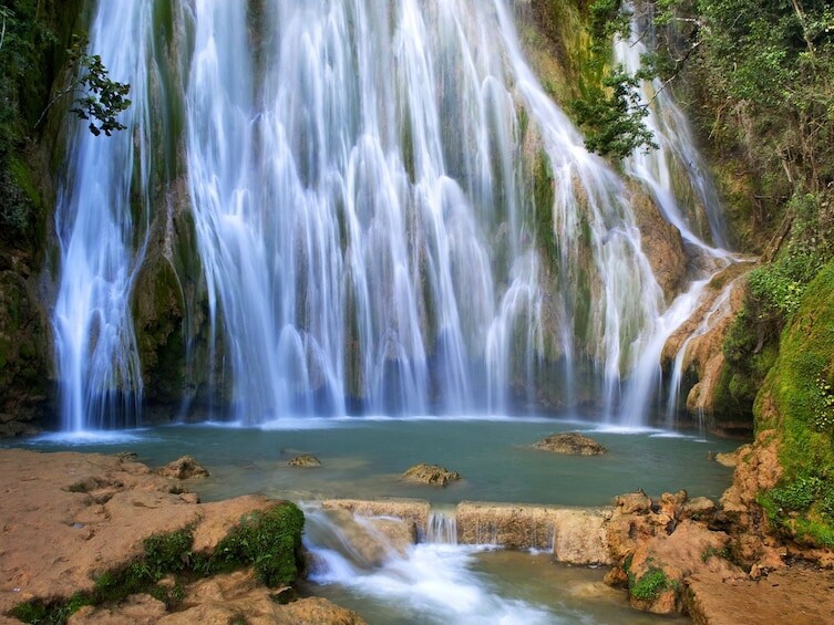 A cascading waterfall is surrounded by lush greenery, flowing over rocky surfaces into a serene pool below. Clear water reflects the surrounding nature, creating a tranquil scene. Moss and vegetation are visible along the rocks, enhancing the natural beauty.