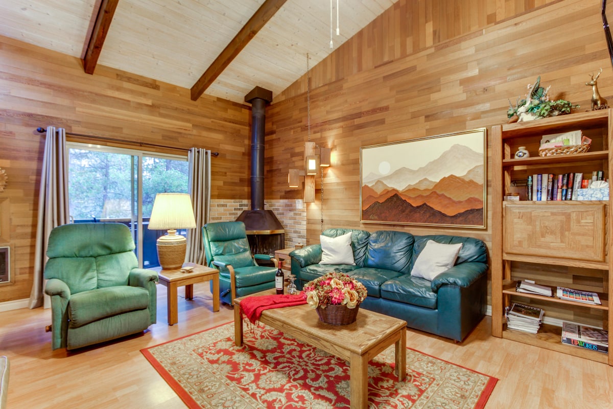 A cozy living area features a wooden wall and ceiling, offering a warm ambiance. Two green leather armchairs and a blue sofa are arranged around a wooden coffee table. A large lamp provides lighting, while a fireplace and shelves with books add to the comfort.