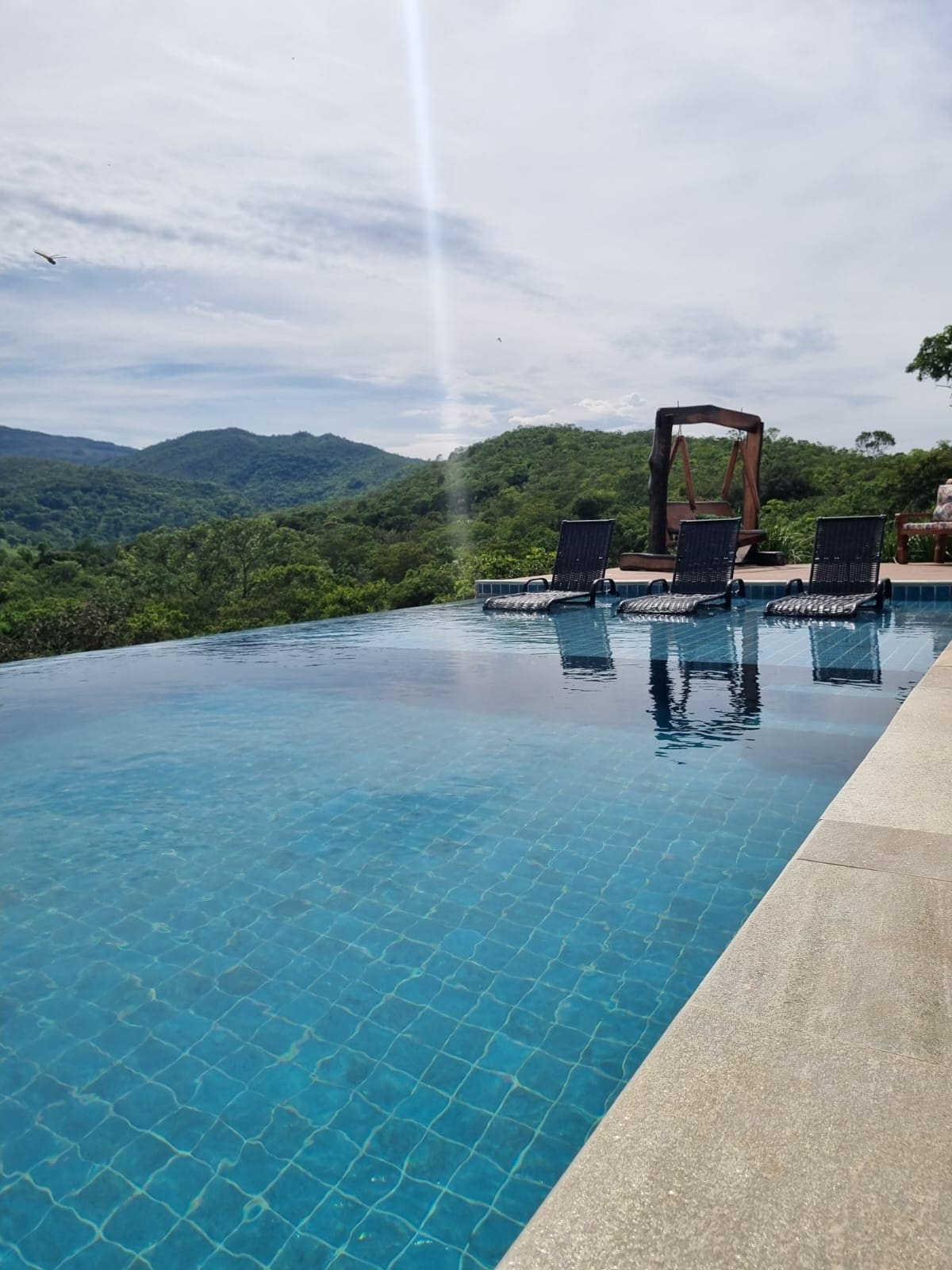 An infinity pool extends towards vibrant green mountains, reflecting the sky's hues. Several lounge chairs are positioned along the pool's edge, inviting relaxation while enjoying the scenic views. Trees frame the background, enhancing the natural surroundings.