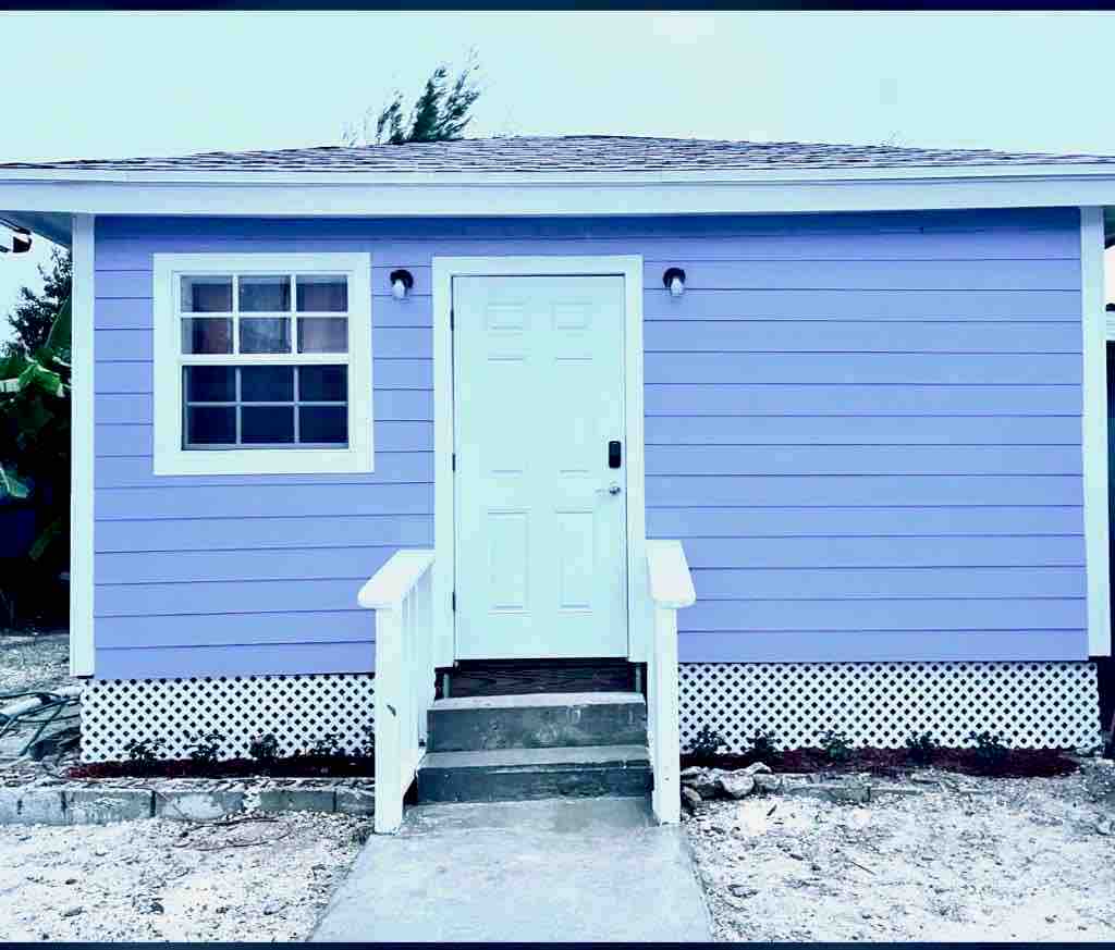 A charming exterior view of a small bungalow, featuring a light purple facade. A white door is centered, flanked by a window on one side. A concrete pathway leads to the entrance, with white steps providing access.