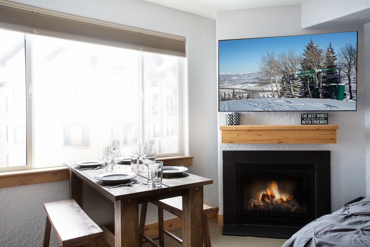 A dining area features a wooden table set for six, with plates and glasses arranged neatly. A large window allows natural light to fill the space, showcasing mountain views. A modern TV is mounted above a cozy fireplace, creating a relaxed ambiance.