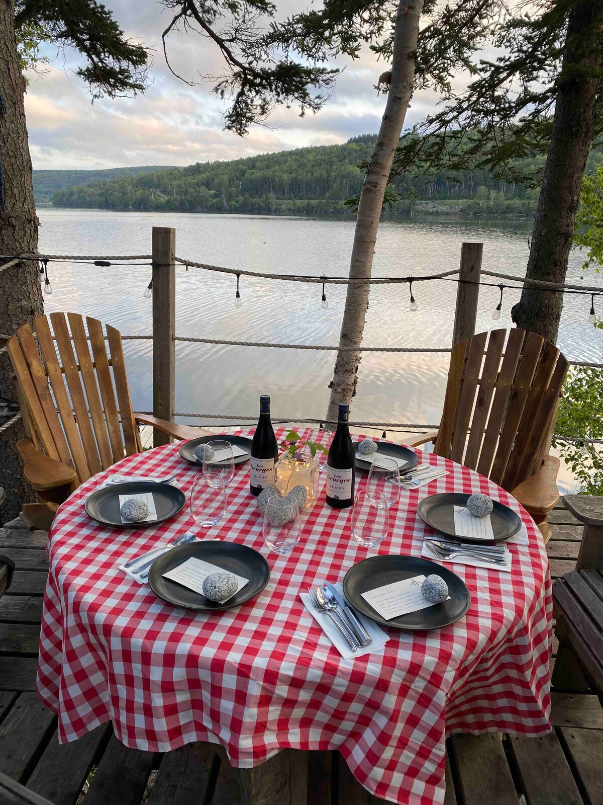 A round table is set with a red and white checkered tablecloth on a deck overlooking the water. Two bottles of wine stand alongside six place settings, each featuring black plates and rolled napkins. Wooden chairs are positioned around the table, with trees providing natural framing.
