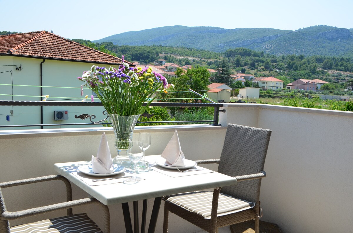 A small balcony features a table set for two. White tableware is arranged neatly alongside crystal glasses. A vase filled with colorful flowers adds a natural touch, while mountain scenery and rooftops are visible in the background, creating a serene outdoor dining space.