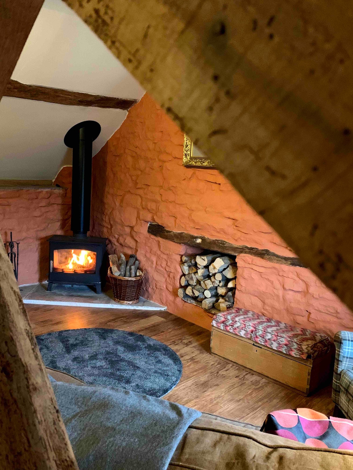 A cozy sitting area features a wood-burning stove with a warm flame, set against an orange wall. A stack of firewood is neatly arranged beside the stove, and a textured rug lies on the wooden floor. A bench with a patterned cushion adds to the inviting ambiance.