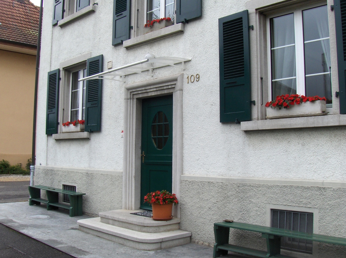 The entrance of a two-story building is framed by green shutters and window boxes with red floral arrangements. A rounded doorway sits atop a small stone step, while two green benches are positioned on either side of the entrance. A small planter enhances the inviting look.