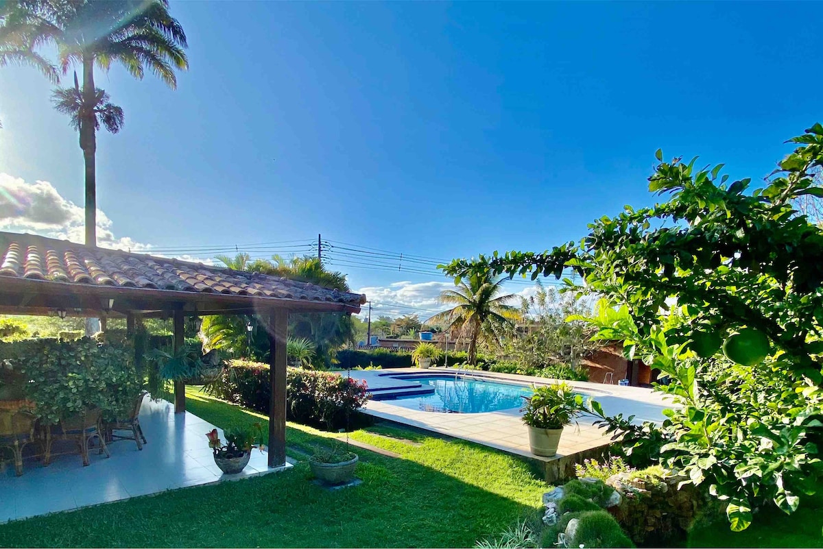 A serene outdoor area offers a view of a swimming pool surrounded by lush greenery. A covered terrace with comfortable seating is visible in the foreground, while a garden is adorned with flowering plants, set against a clear blue sky.