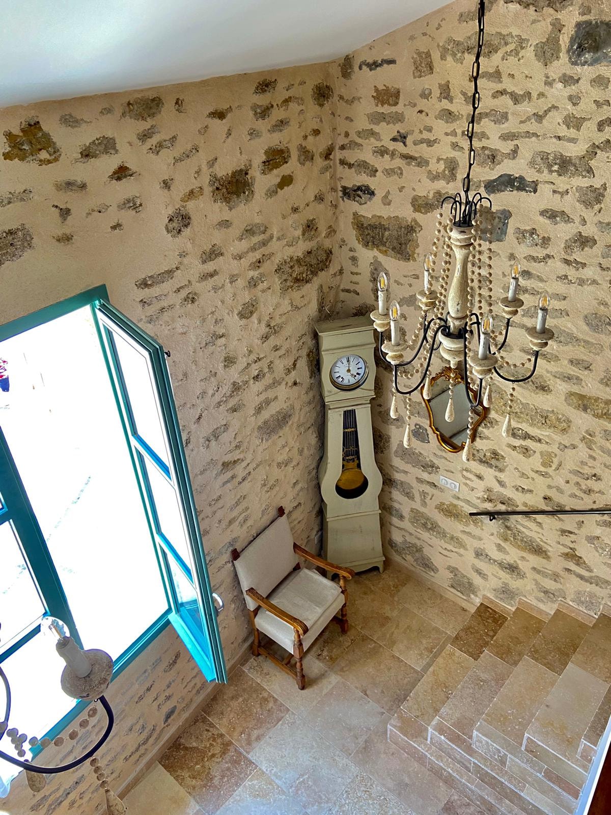 A well-lit stairwell features a large window with turquoise frames, allowing natural light to fill the space. A vintage clock stands beside a minimalist armchair, and an elegant chandelier hangs from the ceiling, complementing the stone walls and stairway leading downward.