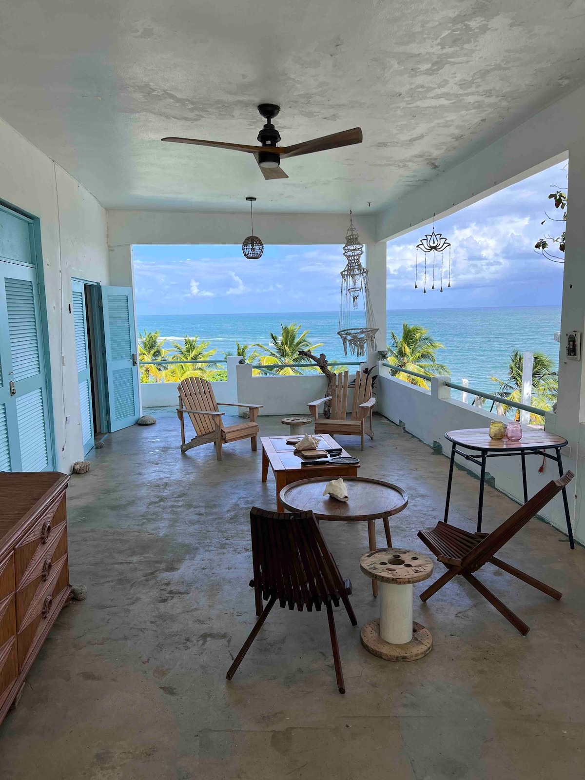 A spacious balcony is furnished with several wooden chairs and a small table, offering a view of the ocean and palm trees. Light colors dominate the décor, complemented by a ceiling fan and decorative elements like wind chimes, enhancing the relaxed atmosphere.