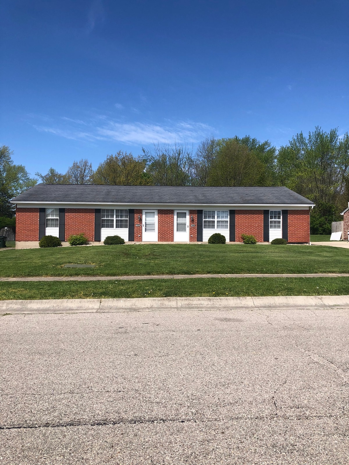 A single-story brick home is presented, featuring a symmetrical façade with three white front doors and six evenly spaced windows. Lush green shrubs flank the entrance, and a clear blue sky is visible overhead, with trees lining the background.