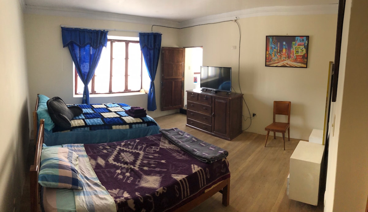 A spacious room is furnished with two beds, one draped in a vibrant blanket. A large window is adorned with blue curtains, allowing natural light to fill the space. A television is mounted on a wooden dresser, complemented by a wooden chair and a nightstand.