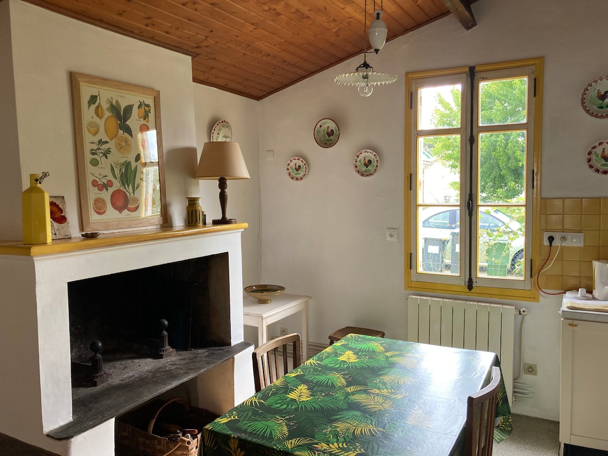 A kitchen area features a table with a tropical print tablecloth, surrounded by wooden chairs. A decorative lamp stands on the table, and a fireplace is visible with a simple design. Yellow accents are evident in the window frames and wall decor showcasing fruit themes.