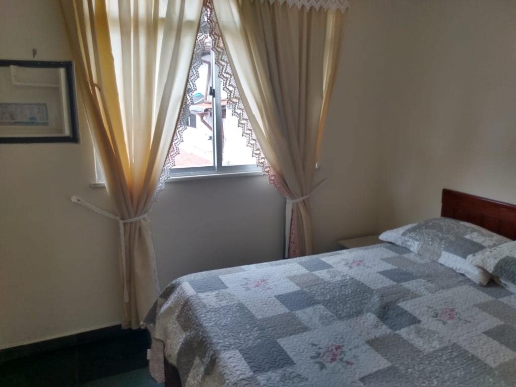 A bright and airy sleeping area features a spacious bed with a patterned quilt. Natural light streams in through a window dressed in light curtains, illuminating the neutral-colored walls and providing a cozy ambiance.