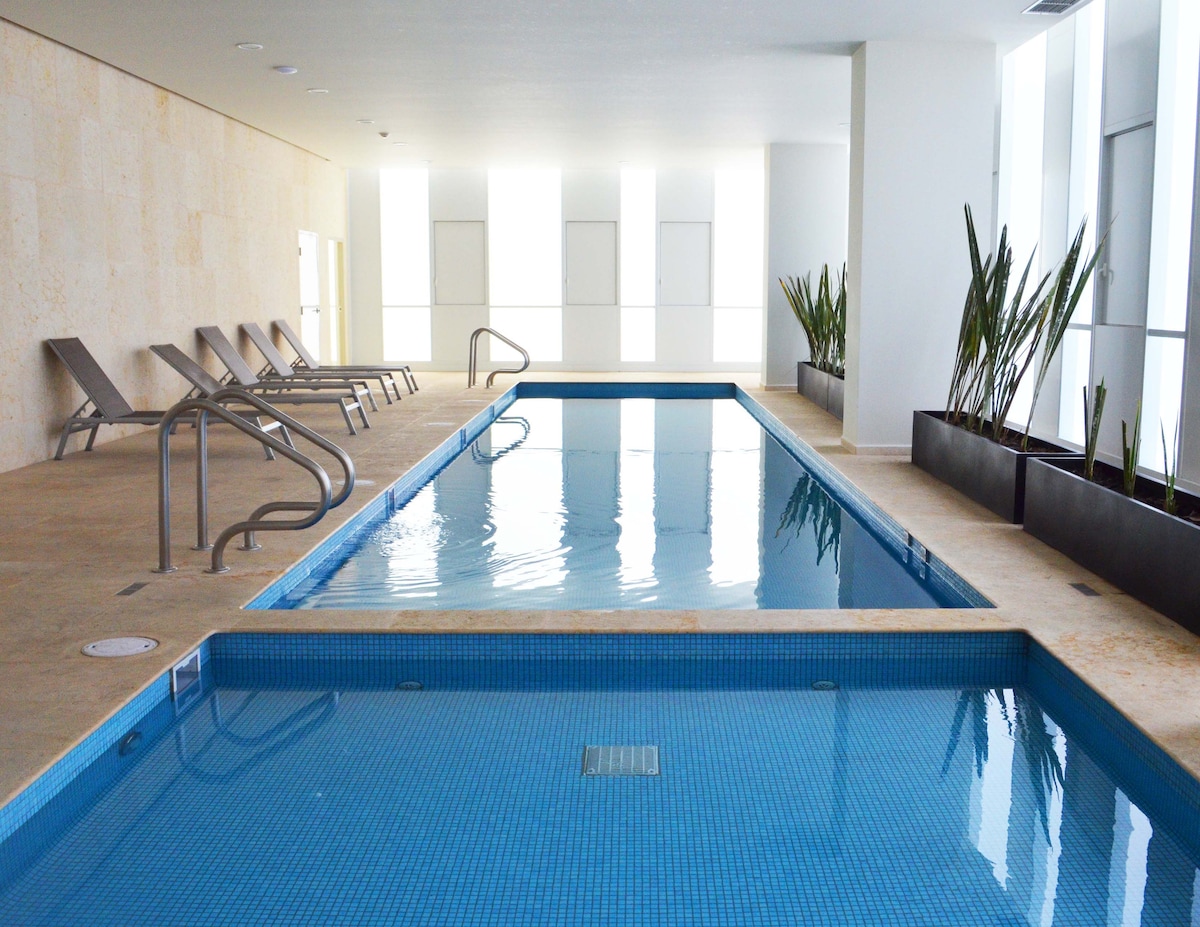An indoor pool area is visible, featuring two swimming pools with clear blue water. Lounge chairs are positioned along the sides, and tall plants are arranged in modern planters along the walls. Natural light floods the space through large windows.