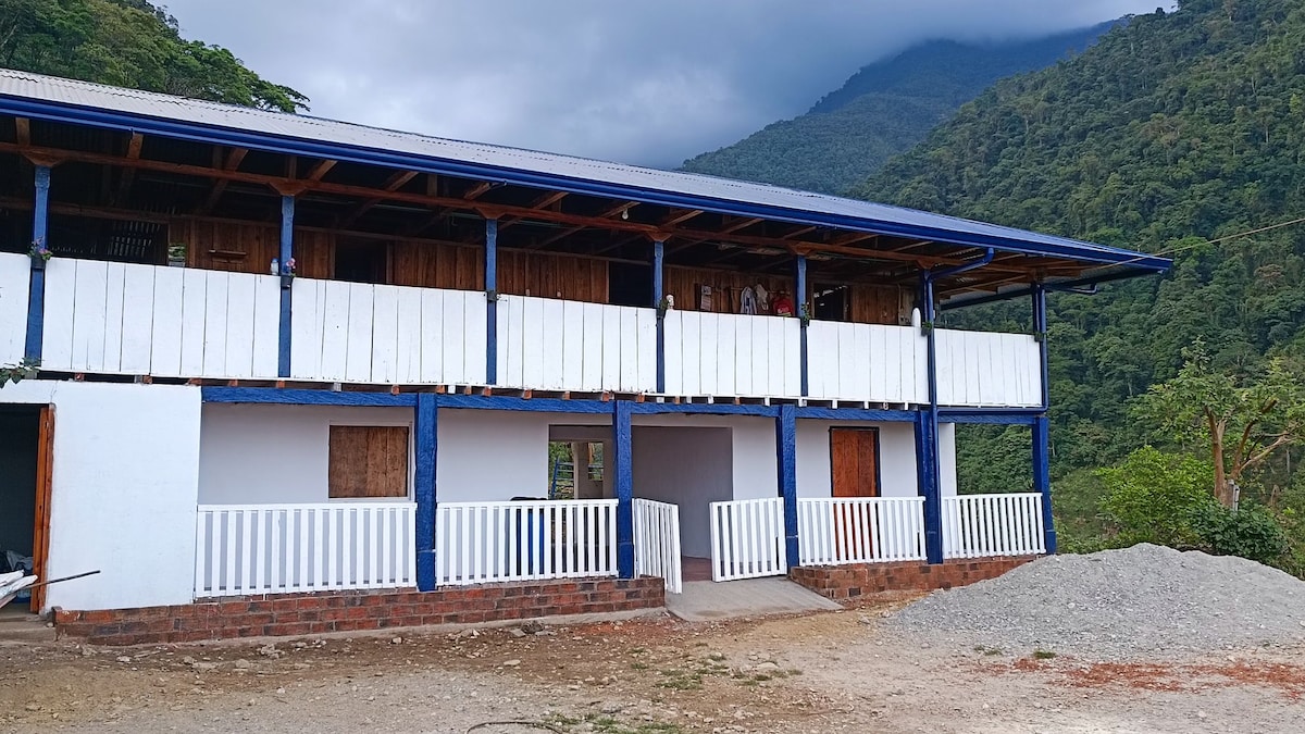 A two-level wooden building is shown with a blue roof and white walls. The structure features multiple balconies with wooden railings, overlooking a lush green mountainous landscape. A gravel area is visible in the foreground, contributing to the rural setting.