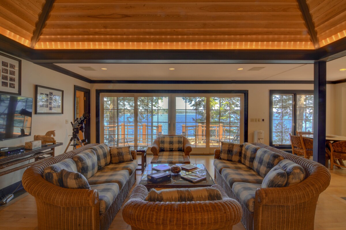 A spacious living area features a series of comfortable wicker sofas arranged around a central coffee table. Large glass doors provide a view of the lake outside, framed by trees and natural light. A telescope is positioned near the windows, enhancing the view.
