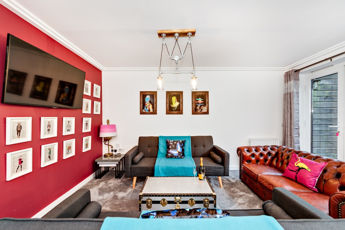 A modern lounge area features a large wall-mounted TV and eclectic artwork displayed in framed prints. A leather chesterfield sofa and two dark sofas provide ample seating, complemented by a stylish coffee table. Natural light streams through the glass doors leading to an outdoor space.