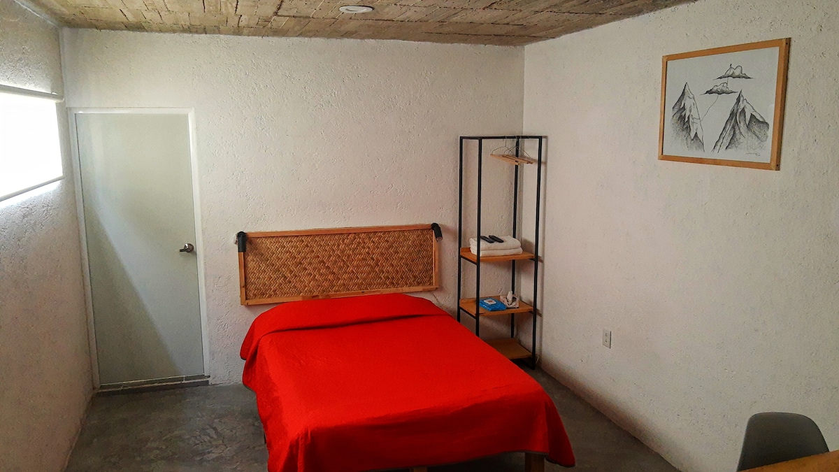A bright room features a cozy bed with a red blanket and a decorative headboard. A wooden shelf holds personal items, and a piece of mountain-themed artwork adorns the wall. Natural light enters through a window, complementing the neutral colored walls and gray flooring.