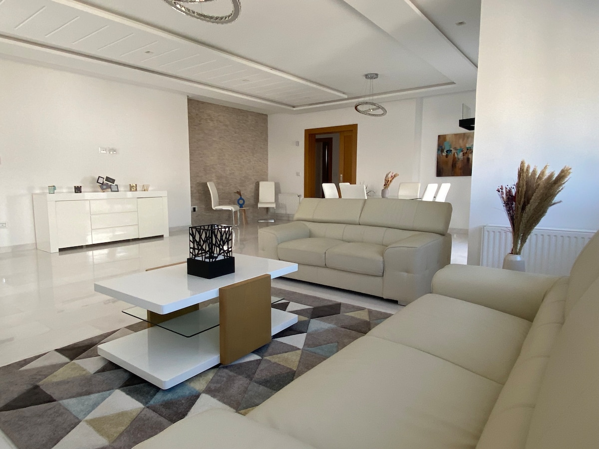 A modern living area is presented with light-colored leather sofas arranged around a geometric-patterned rug. A chic coffee table features a minimalist design in the center, with a decorative centerpiece. Neutral wall tones and bright lighting create an open and airy ambiance.