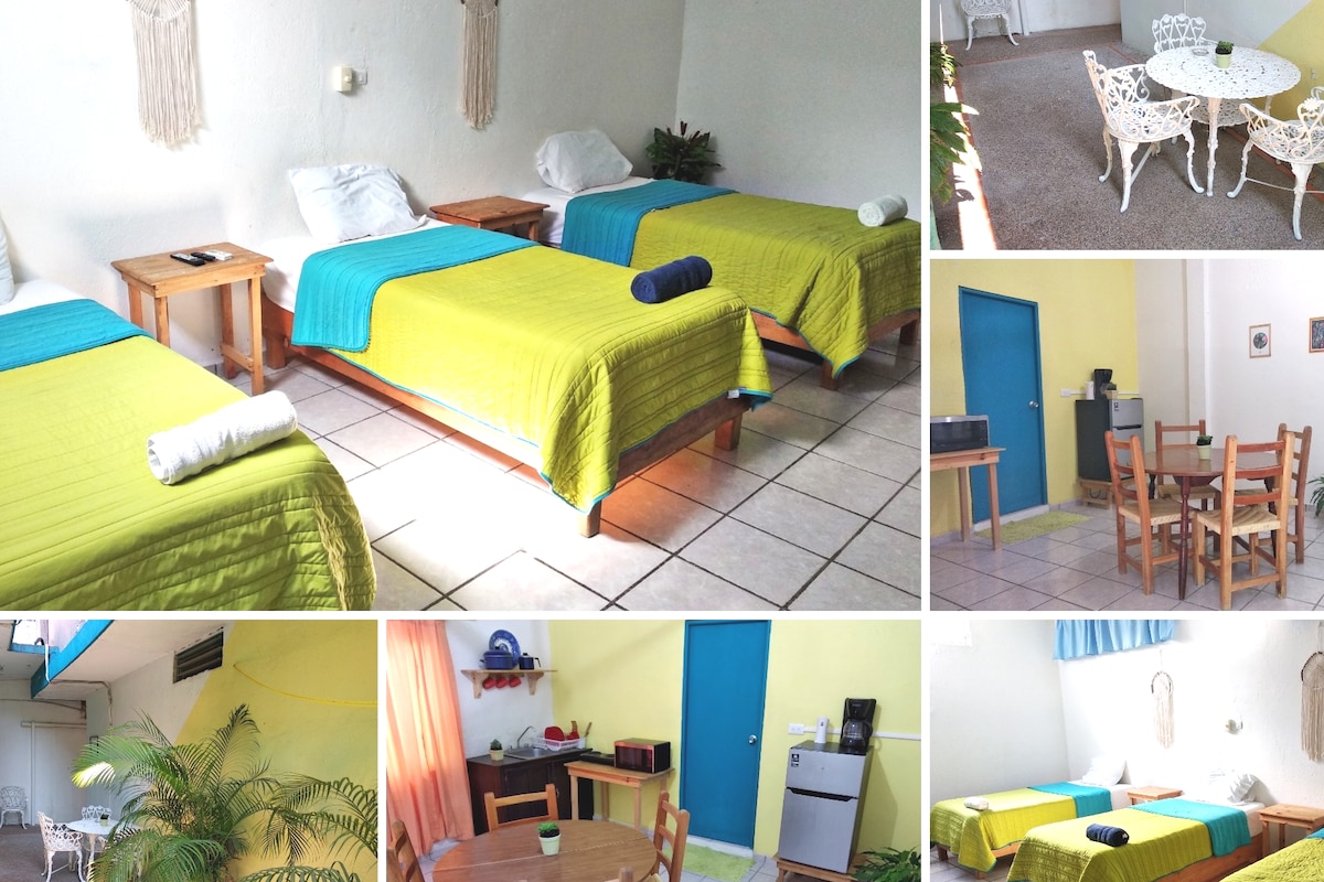 A well-lit room features three individual beds covered in vibrant yellow and blue linens, providing a spacious and inviting sleeping area. Small wooden nightstands hold fresh towels, while a dining table with white wicker chairs invites meals. A kitchenette area with a microwave and refrigerator is visible.