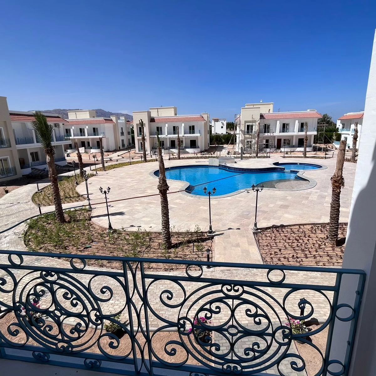 A view from a balcony overlooking a spacious pool area, surrounded by landscaped pathways and palm trees. The pool features a unique shape and is bordered by lounge chairs. Nearby buildings are visible, creating an inviting communal atmosphere.