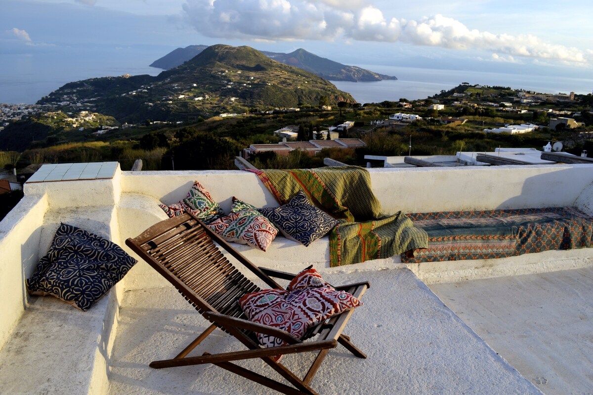 A spacious outdoor patio features a comfortable folding chair and a cushioned bench, adorned with patterned cushions. The area offers panoramic views of rolling hills and the sea, capturing the serene beauty of the surrounding landscape.