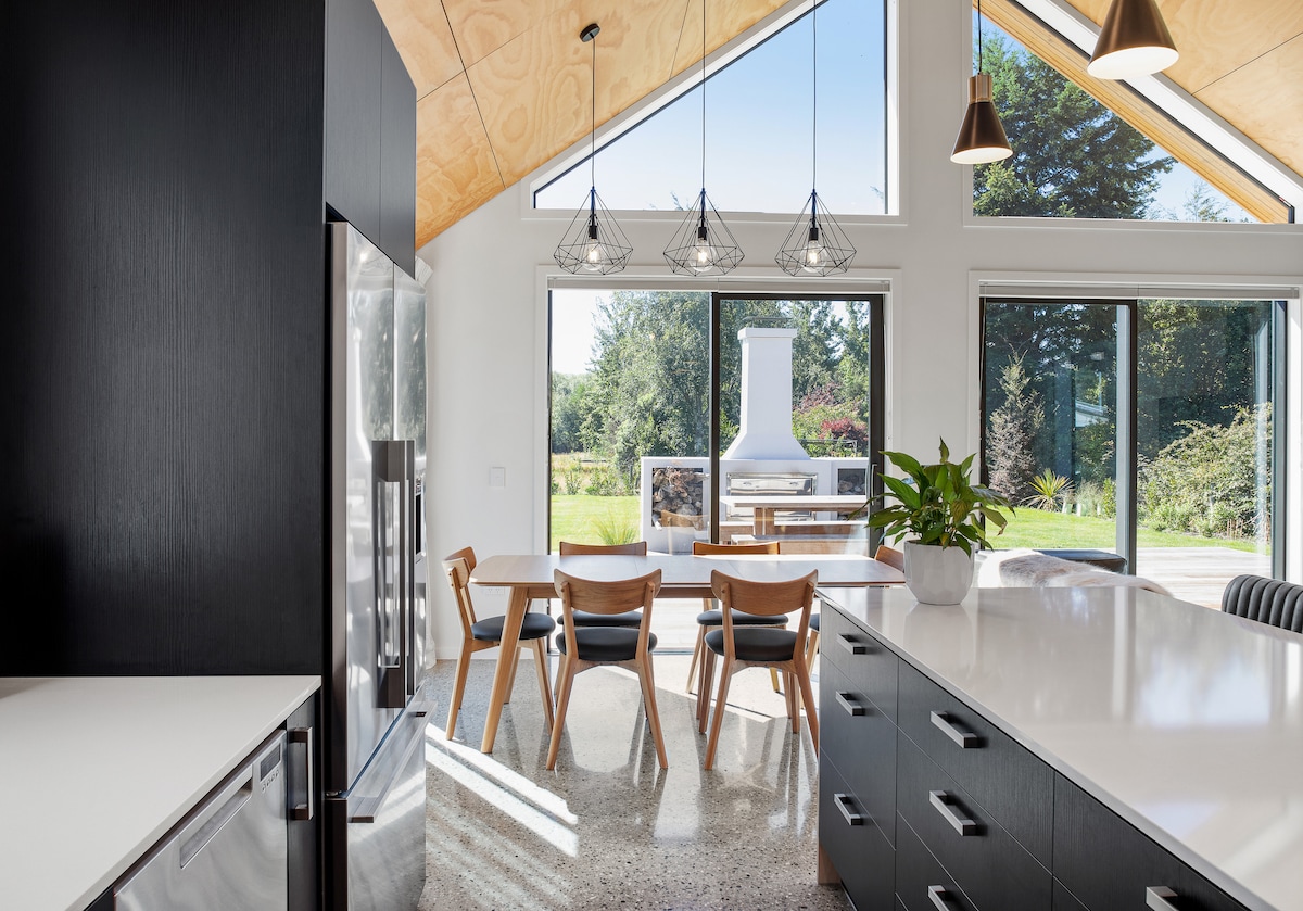 A modern kitchen is featured, showcasing a large island with a white countertop. Natural light floods the space through expansive windows, illuminating the dining area with wooden chairs. Minimalistic pendant lights hang from a high ceiling, enhancing the open and airy environment.