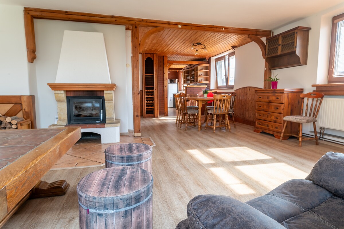 A spacious living area features a wooden dining table surrounded by chairs, alongside a cozy seating arrangement. A fireplace sits within a stone surround, with a wooden storage area for logs nearby. Natural light filters in through windows, enhancing the warm ambiance.