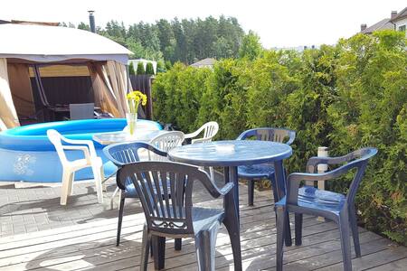 A wooden deck is equipped with several outdoor seating options, including a round table surrounded by four chairs. An inflatable pool is positioned nearby, with lush greenery providing a natural border. A bright vase of flowers is placed on the table, adding a touch of color.