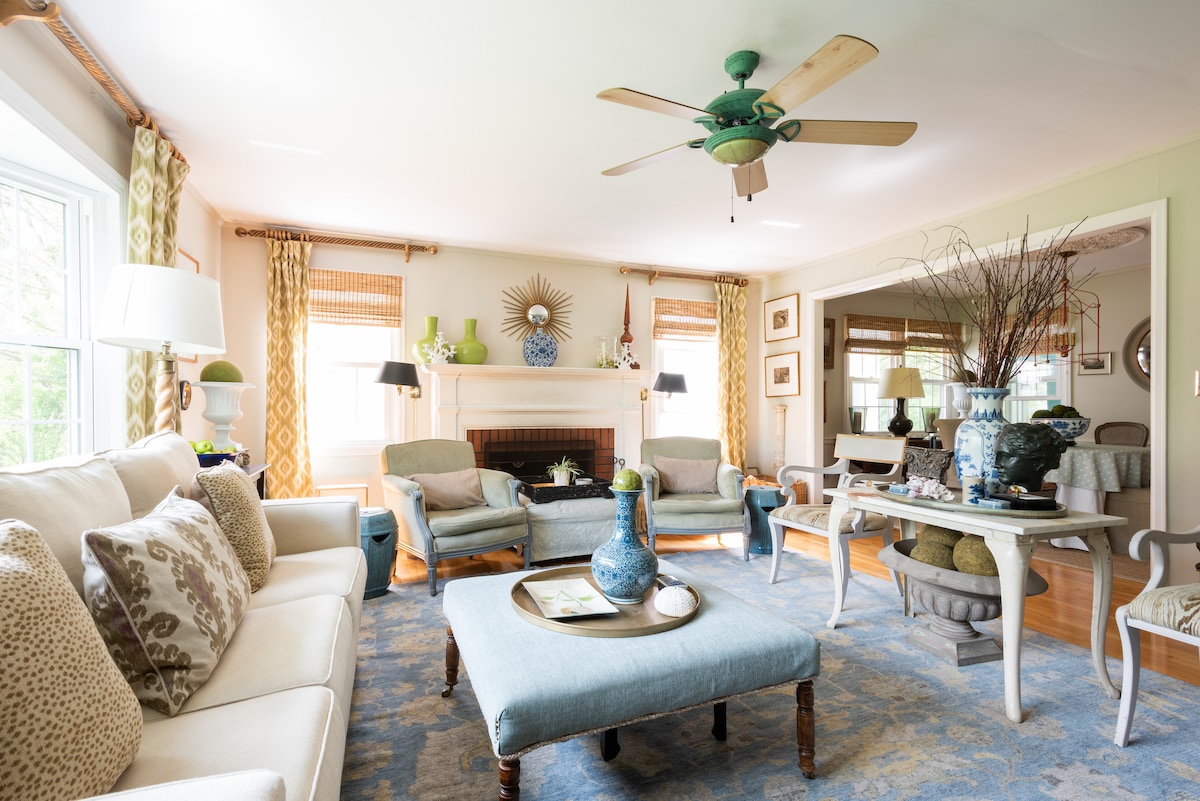 A cozy living room features a comfortable beige sofa and matching armchairs, accented by decorative pillows. A central coffee table holds a vase, and light filters through the large windows adorned with cheerful curtains. A fireplace is framed by a stylish mirror, enhancing the inviting atmosphere.