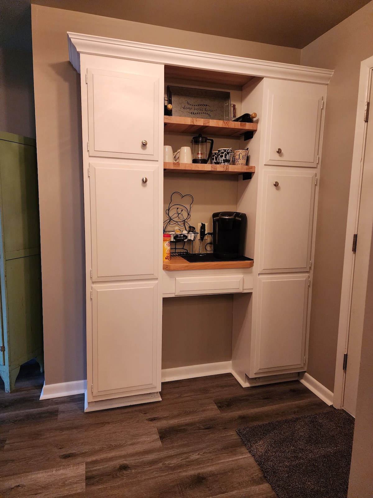 A white cabinet with multiple doors features open shelving above, showcasing a coffee maker and various kitchen items. A small, wooden countertop is available beneath the shelves, contributing to a functional kitchen space.