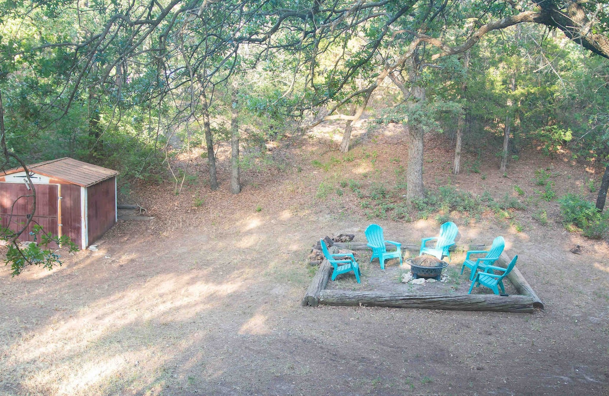 A designated fire pit area is surrounded by six turquoise chairs, arranged in a circle on a natural ground surface. A wooden log serves as a border, and a small, rustic shed is partially visible in the background among the trees.