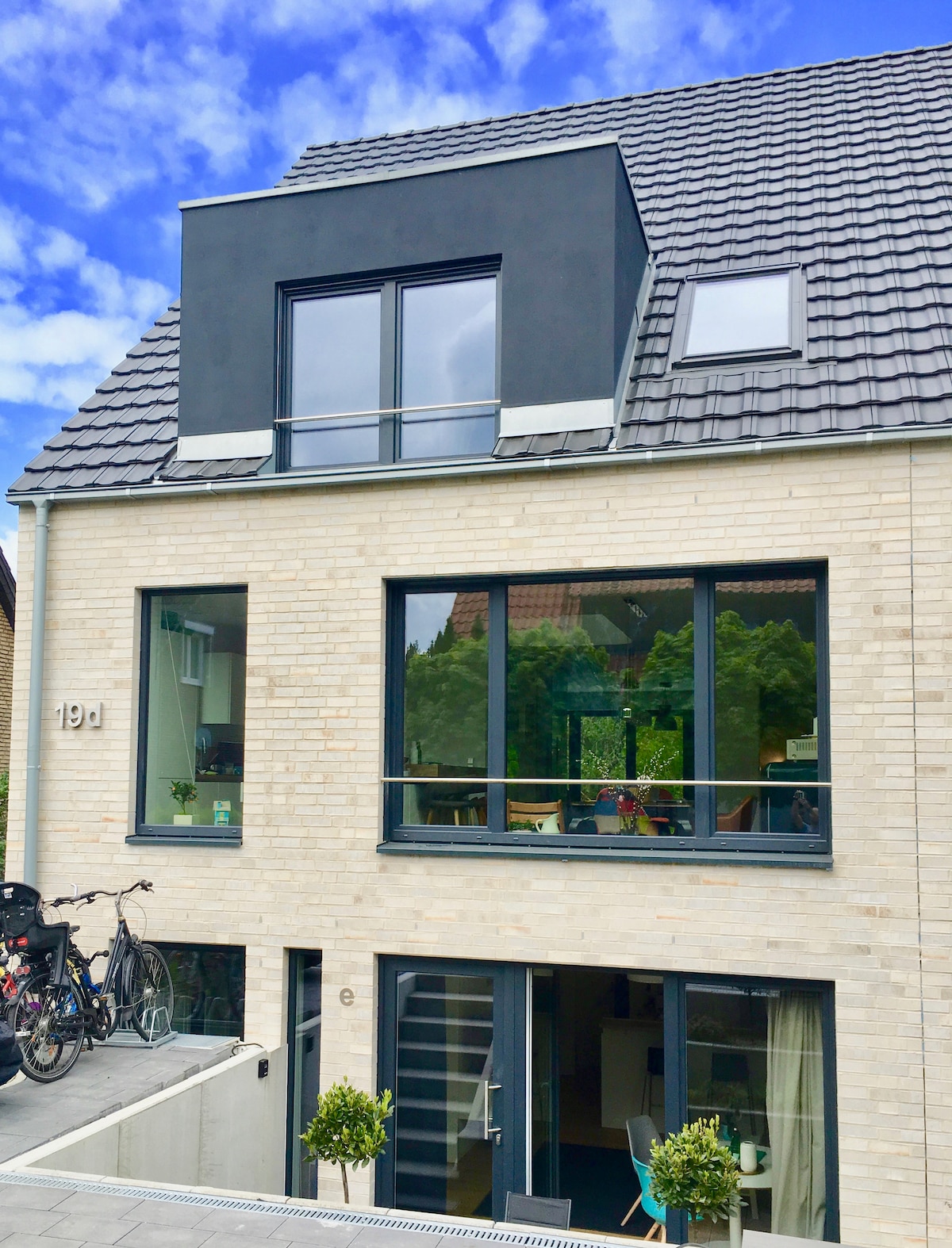 A modern building features a sleek grey facade and large windows, allowing for ample natural light. A set of bicycles is parked outside, and potted plants add a touch of greenery. The entrance is marked by a small staircase leading to the main door.