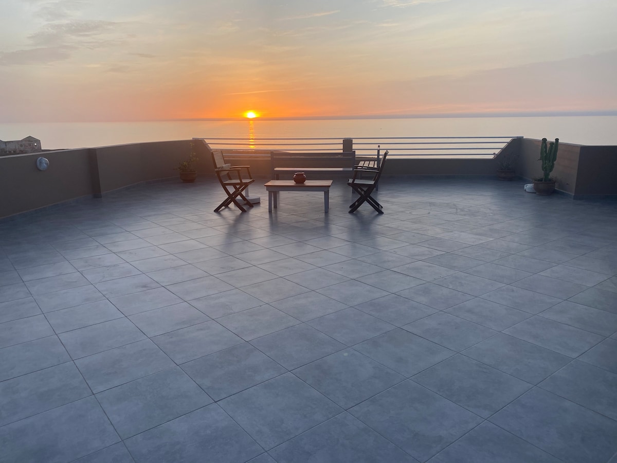 A spacious balcony features tiled flooring and a clear view of the sunset over the ocean. Two wooden chairs are positioned alongside a small table, inviting relaxation. Subtle greenery is present in the form of potted plants, enhancing the outdoor space.