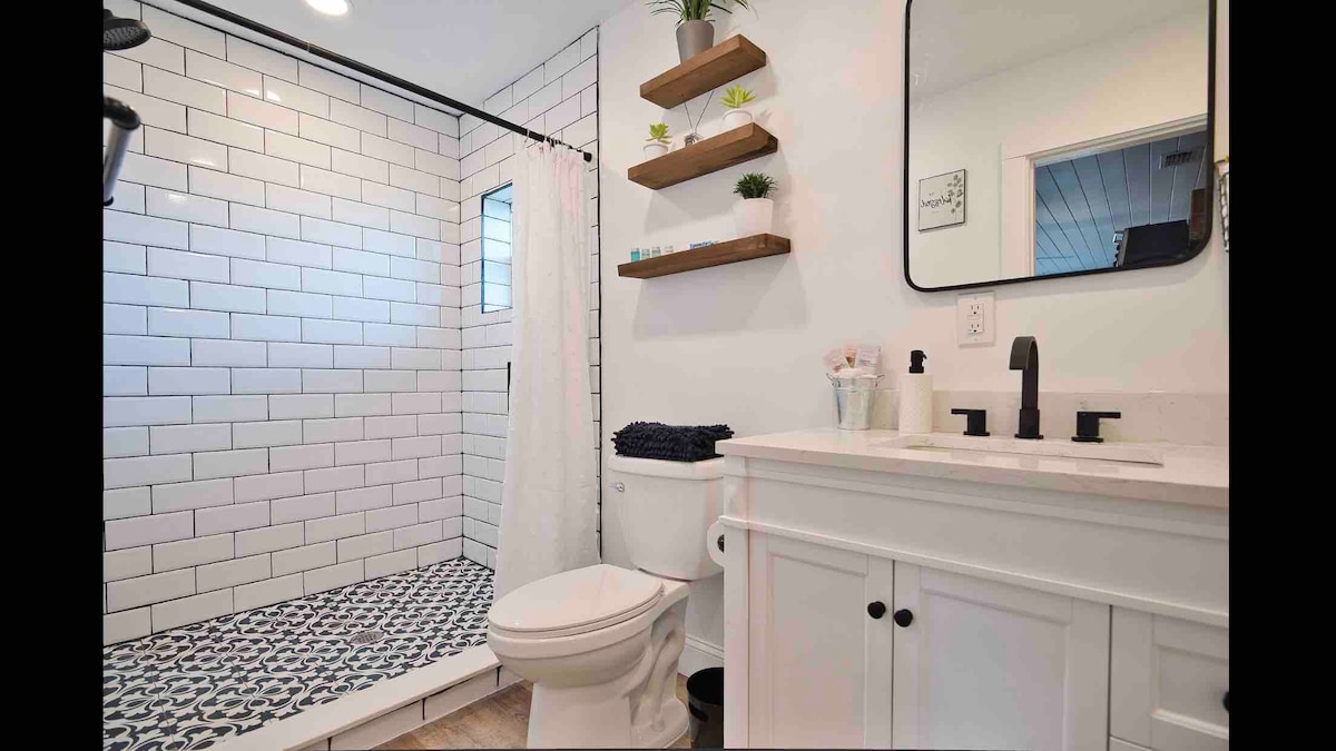 A modern bathroom features a spacious shower with white tiled walls and a patterned floor. Shelves hold small plants and decorative items. A white vanity with two sinks complements a clean design, while a large mirror enhances natural light.