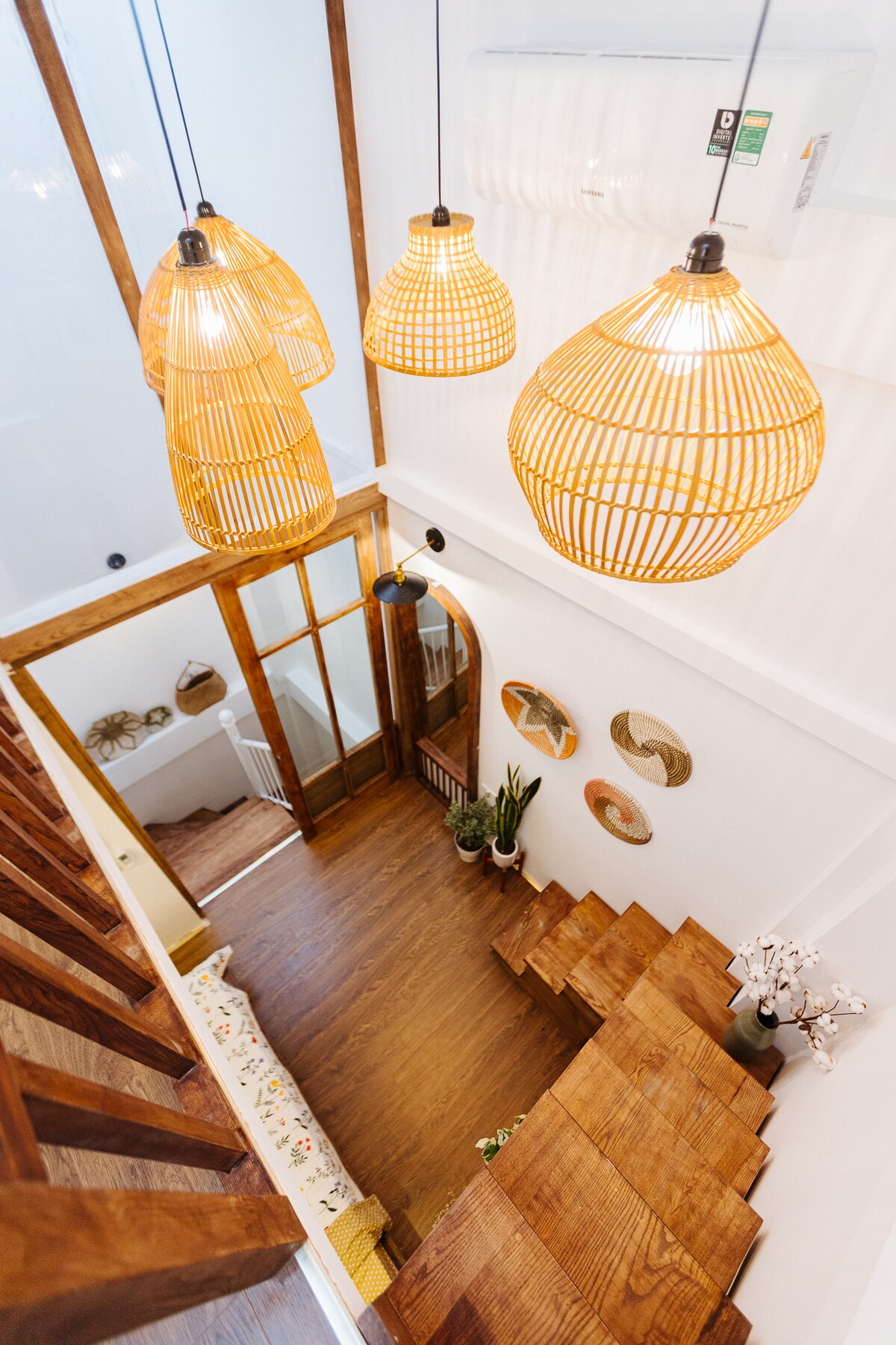 A view from above captures the warm ambiance of the home, showcasing three woven pendant lights hanging gracefully. Wooden stairs lead down to the lower floor, bordered by decorative wall art. A cozy seating area with cushions is visible, enhancing the inviting environment.