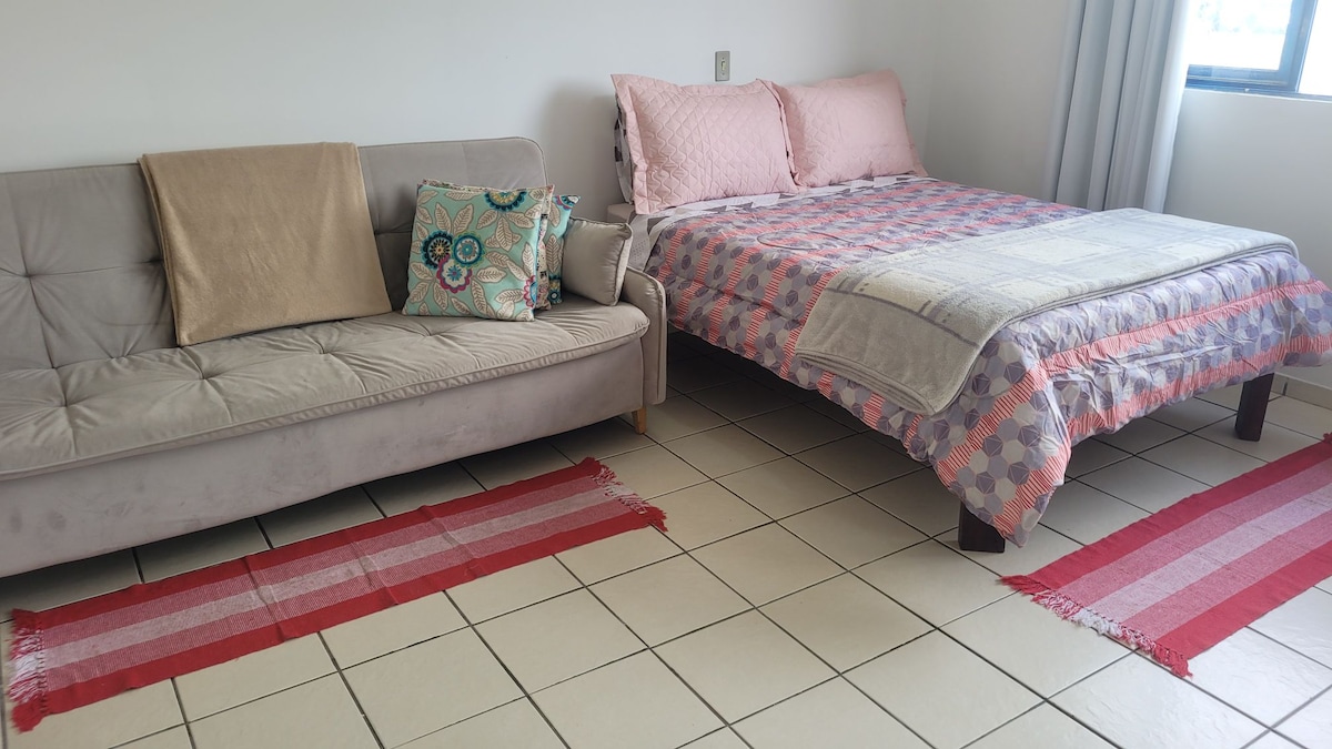 A sleeping area is illustrated with a spacious bed dressed in a patterned quilt, flanked by two decorative pillow sets. A sofa bed is positioned nearby, complemented by a decorative cushion. Area rugs add warmth to the tiled floor, enhancing the overall comfort of the space.